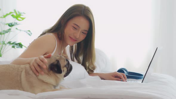 Woman typing and working on laptop with dog Pug breed lying on bed alt