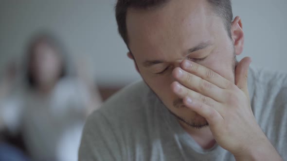 Close Up Portrait of Crying Man on the Foreground and His Shouting Angry Wife Sitting on the Sofa alt