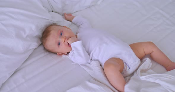 Pretty Baby with Big Blue Eyes is Lying on a White Bed and Sucking His Fingers alt