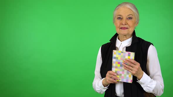An elderly woman holds a book, smiles at it and at the camera - green screen studio alt