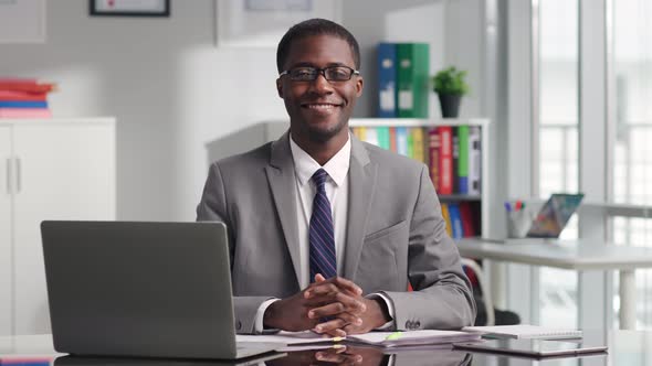 Portrait of AfricanAmerican Politician in Glasses Smiling at Camera Sitting at Desk alt