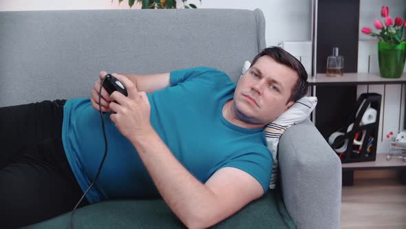 A man lies on the couch in front of the TV in his hands holding a joystick, plays the console