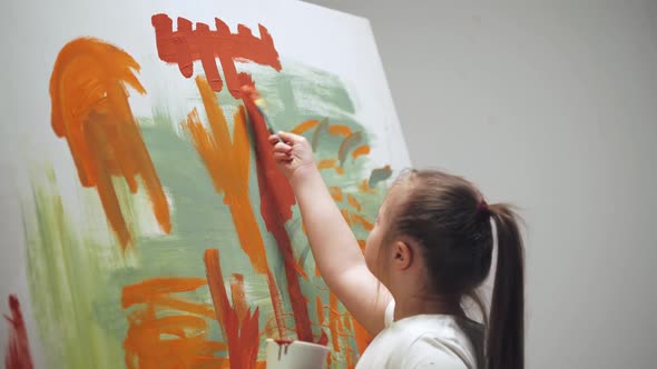 Kid Girl with Down Syndrome Draws with a Brush on a Large Canvas in a White Room Girl with Special alt