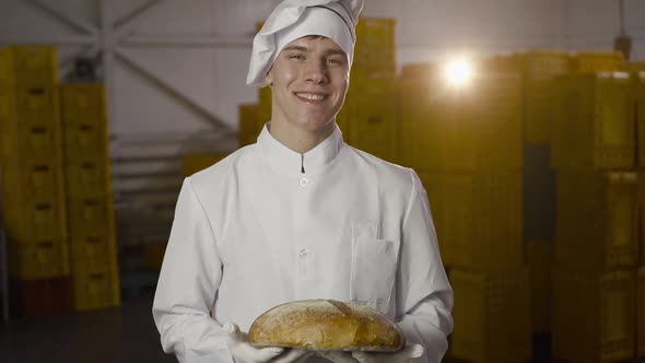 Happy Baker in Uniform Holds a Fresh Loaf of a Bread in a Bakery with Backlit alt