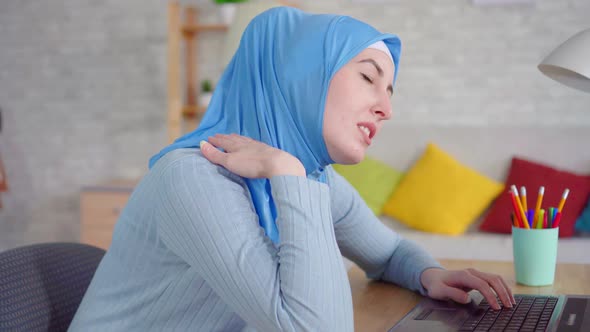 Young Muslim Woman in a Traditional Headscarf Slouches While Sitting at a Laptop and Feels Pain in alt