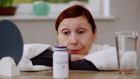 Closeup Thoughtful Mature Caucasian Woman Sitting at Table with Vitamins Bottle alt