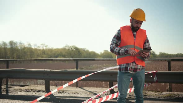Young Bearded Man Inspector in an Orange Vest and Protective Helmet with Tablet alt