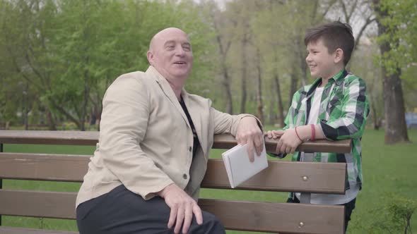 The Bald Mature Man Sitting in the Park on the Bench with a Book, Talking with His Grandson alt