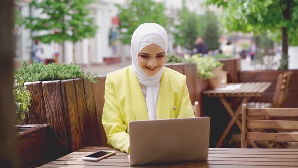 Smiling Young Muslim Woman Wearing Headscarf Sits in Cafe and Uses Laptop