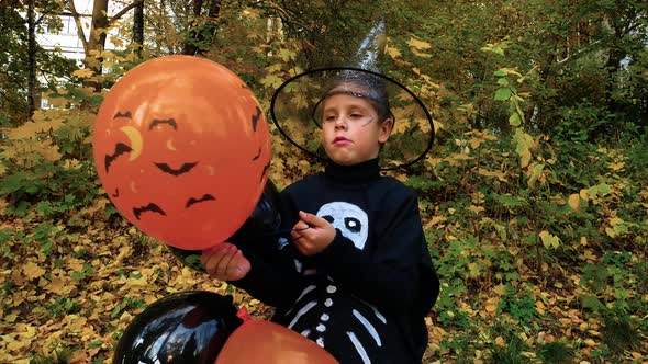 A Boy in a Skeleton Costume and a Wizard's Hat Holds Orange Black Balloons in His Hands