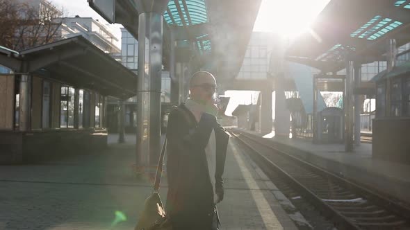 Business Man in a Raincoat and Sunglasses Smokes a Cigarette on the Platform of the Railway Station alt