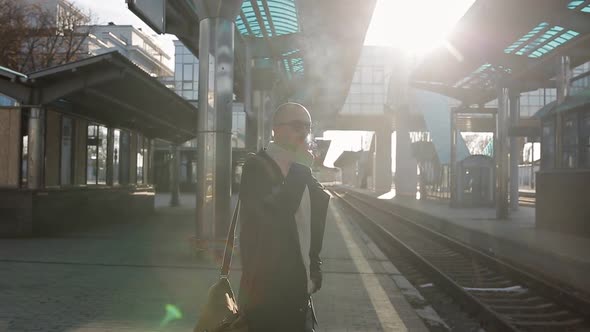Business Man in a Raincoat and Sunglasses Smokes a Cigarette on the Platform of the Railway Station