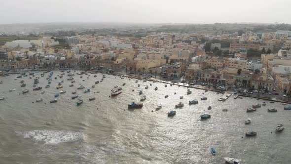Aerial Wide Pan Right Shot Of Marsaxlokk Port With Village Background In Malta alt