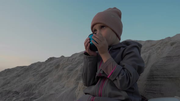 Little girl sitting on a sandy beach and sipping tea alt