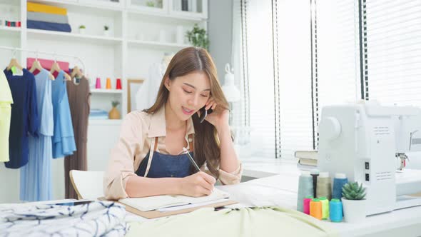 Asian tailor woman talking on phone with customer in tailoring atelier. alt