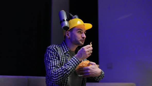 Man With Beer Helmet on the Head Eats Chips While Sitting at Home on Couch