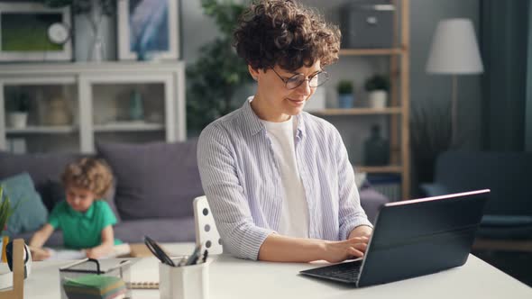 Happy Mother Working at Home with Laptop Typing While Small Boy Son Drawing alt