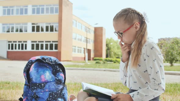 11 Year Old Schoolgirl Reads a Book While Sitting on the Grass on the Grass Near the School. alt