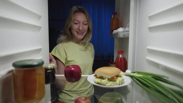Young Woman Opens the Refrigerator Door at Night and Taking Apple Instead of Burger alt