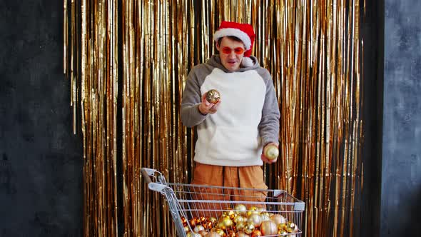 Young Male with Shopping Cart Filled of Assorted Baubles Standing Against Wall with Golden Tinsel alt