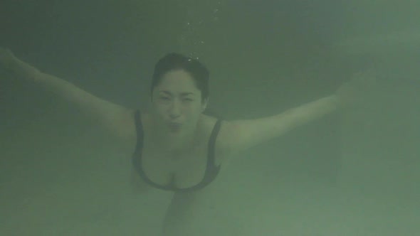 Woman swimming underwater in pool and then emerging to smile at camera