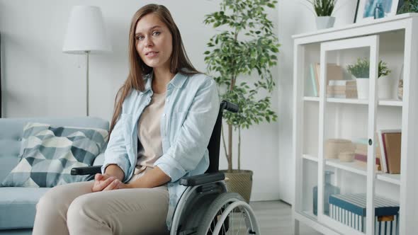 Portrait of Young Disabled Woman Sitting in Wheelchair at Home and Looking at Camera with Light alt