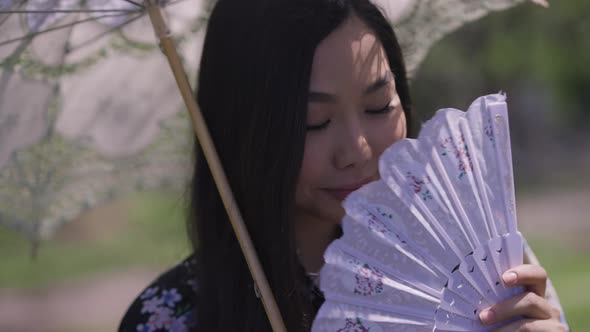 Closeup of Charming Shy Asian Young Woman with White Sun Umbrella Shaking Fan Smiling alt
