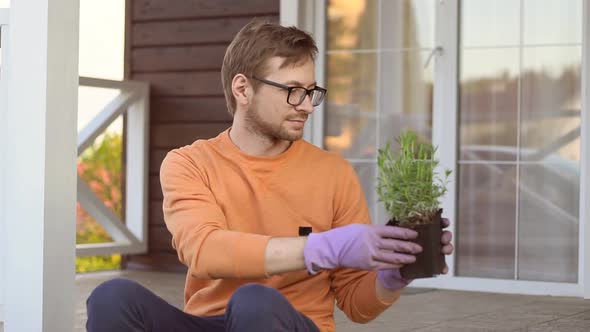 Young man gardener wearing gloves holding a young plant lavender in plastic pot. alt