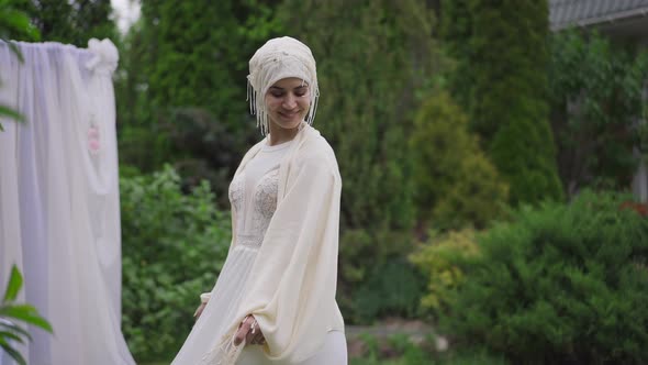 Joyful Smiling Middle Eastern Bride in White Wedding Dress and Hijab Spinning in Summer Garden at alt