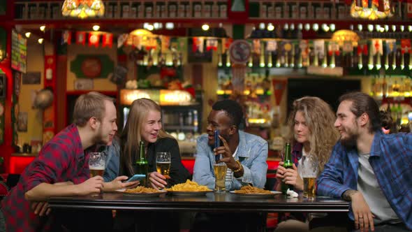 African American Shows Friends Pictures on the Phone They Look and Laugh While Sitting in a Bar in alt
