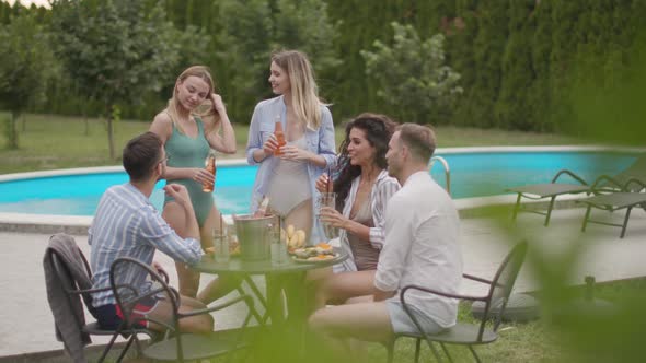 Group of happy young people cheering with drinks and eating fruits by the pool in the garden