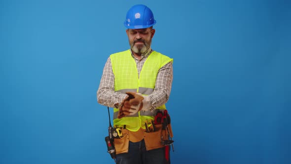 Senior Man Builder Puts on Protective Gloves Against Blue Background