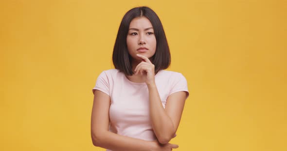 Young Thoughtful Asian Woman Pondering About Something, Touching Her Chin, Orange Studio Background alt