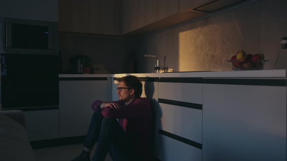 Young man with a thoughtful look sits on the kitchen floor at home and looks ahead alt