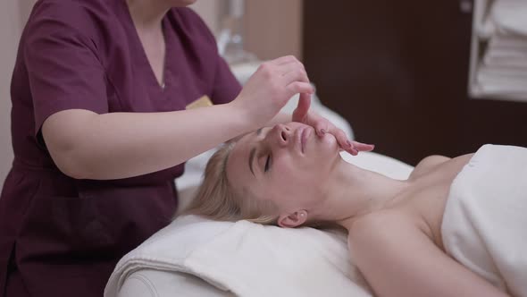 Beautiful Blond Caucasian Woman Enjoying Facial Massage Lying in Spa Salon with Unrecognizable alt