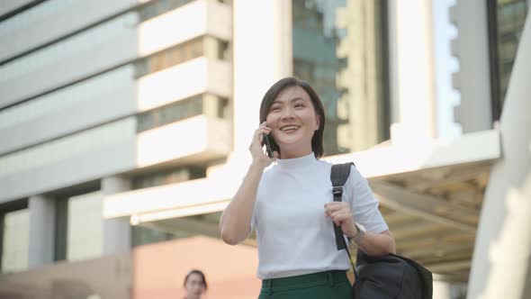 Happy asian woman walks down the city street and talking on mobile phone.