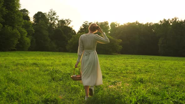 A Woman in a Sundress Walks with a Basket of Fruits on the Grass at Sunset