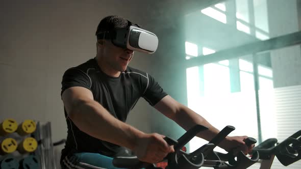 A Man Performs a Heavy Exercise on a Bicycle Simulator in a VR Helmet alt