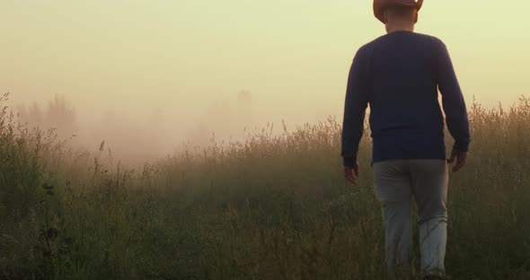 Young Man in Cowboy Hat Walks Through a Field in Fog and Admires the Setting Sun