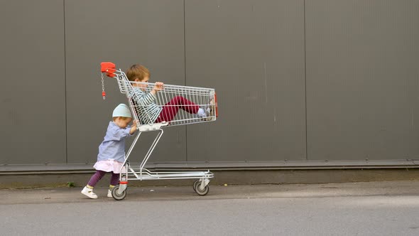 Little Girl Pushing Shopping Cart with Brother Sitting in It. Children Entertainments While Waiting alt