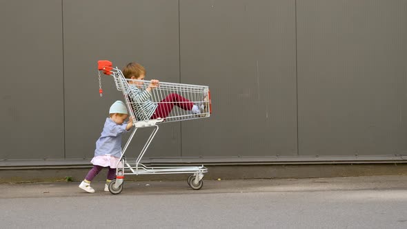 Little Girl Pushing Shopping Cart with Brother Sitting in It. Children Entertainments While Waiting