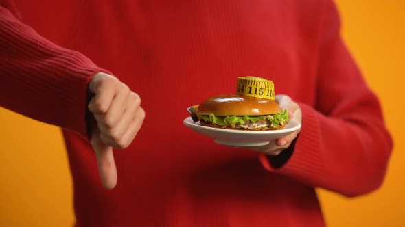 Female Holding Plate With Burger and Measuring Tape Showing Thumbs Down, Diet alt