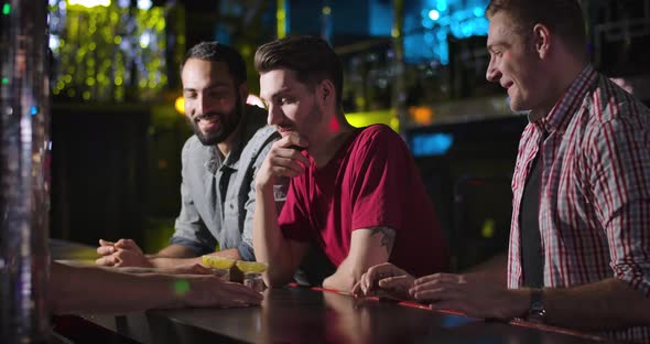 Three Young Men Standing at Bar Counter and Talking with Bartender, Barkeeper Giving Short Drinks alt