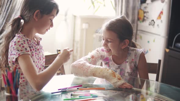 Child with a Broken Arm in a Bandage Laughs and Smiles When Her Sister Paints Her a Cast alt