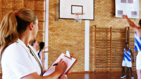 Female coach writing on clipboard while students playing in basketball court alt