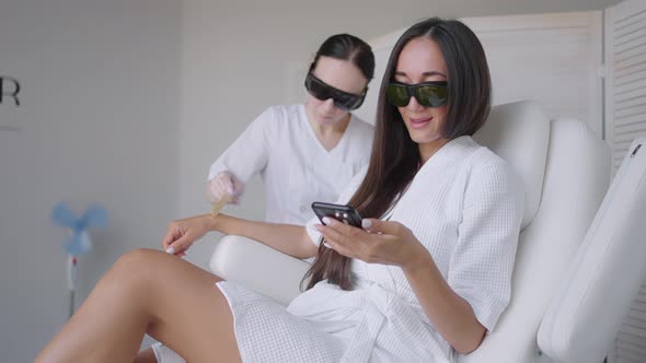 During a Laser Hair Removal Session a Woman in a Beauty Salon Writes a Message in Her Mobile Phone alt