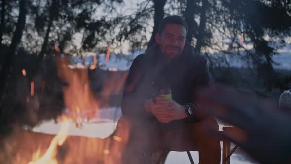 A Young Man in a Jacket Sits By the Fire at Night with a Mug of Hot Tea alt