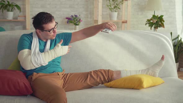 Young Man with a Broken Arm and Leg Sitting on the Couch Talking on Video Call alt