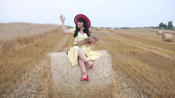 A Beautiful Sweet Brunette Woman in a Field with Hay on a Haystack in a Yellow Stylish Dress