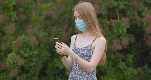 A young girl in a protective medical mask looks at the phone and sends messages